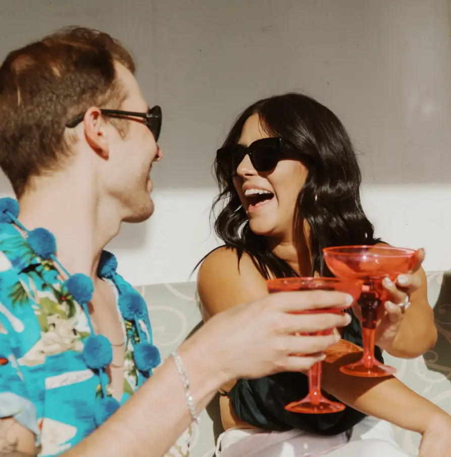 deux personnes souriantes portant des lunettes de soleil trinquent avec des verres à cocktail rouges lors d'une fête estivale