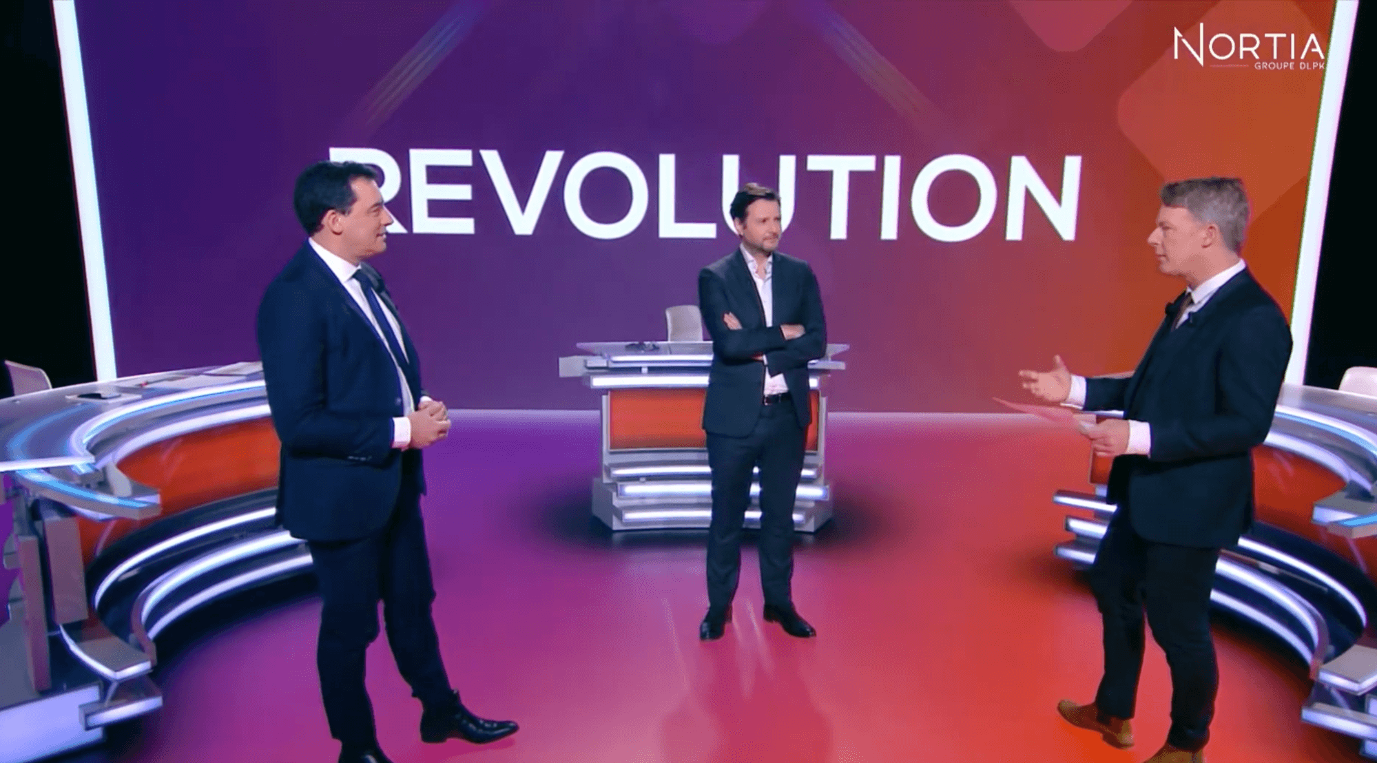 trois hommes en costume discutant sur un plateau de télévision avec le mot 'Révolution' en arrière-plan